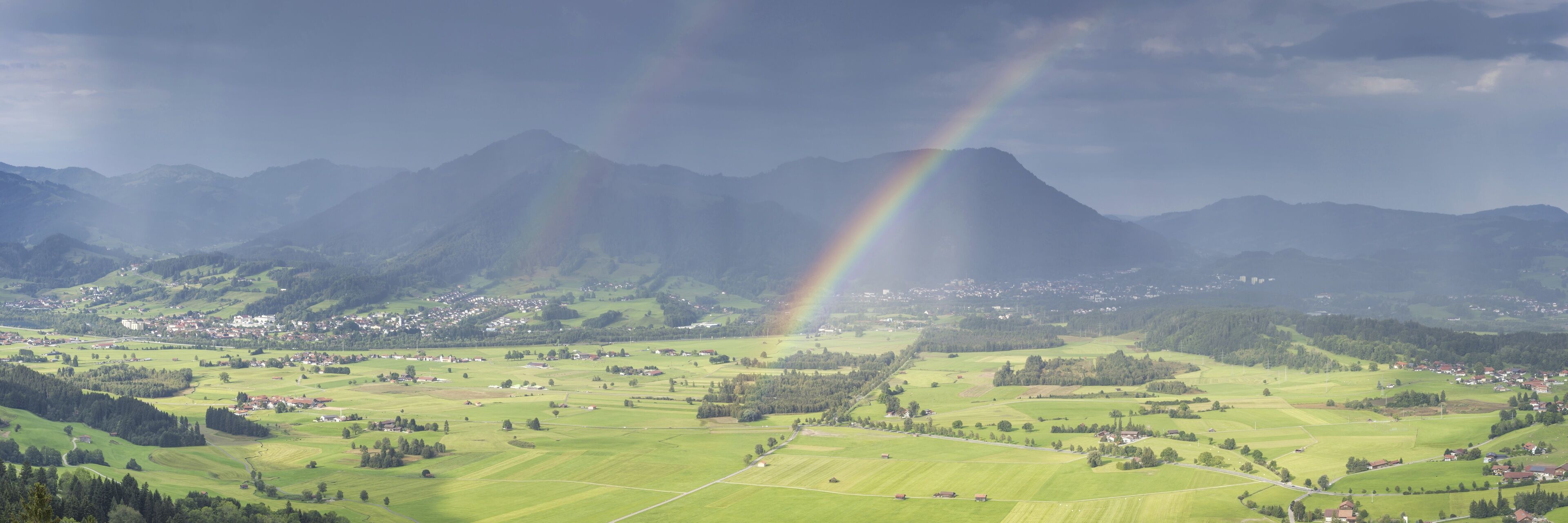 A mountain range with a rainbow over a valley with scattered houses and fields.