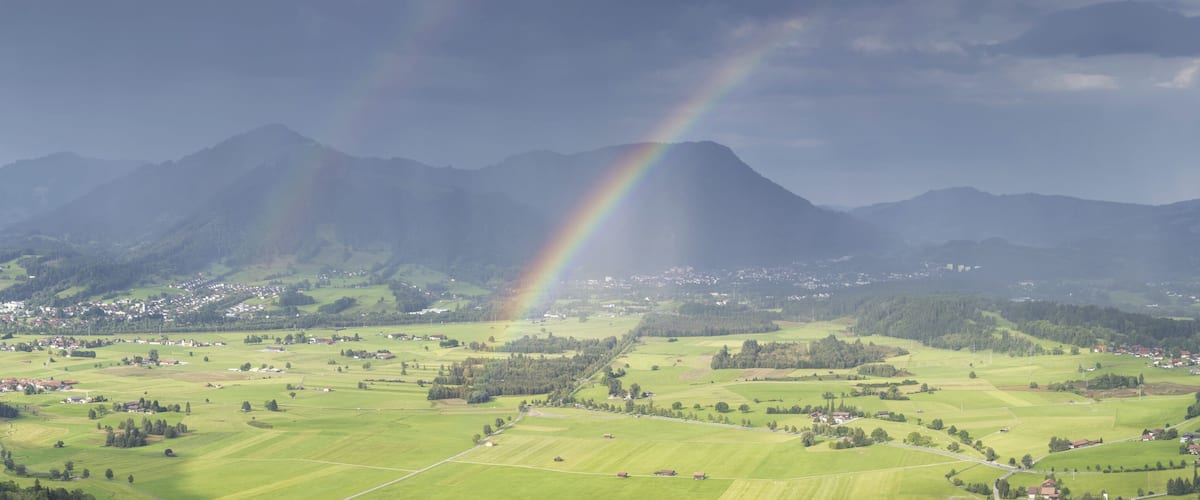 A mountain range with a rainbow over a valley with scattered houses and fields.