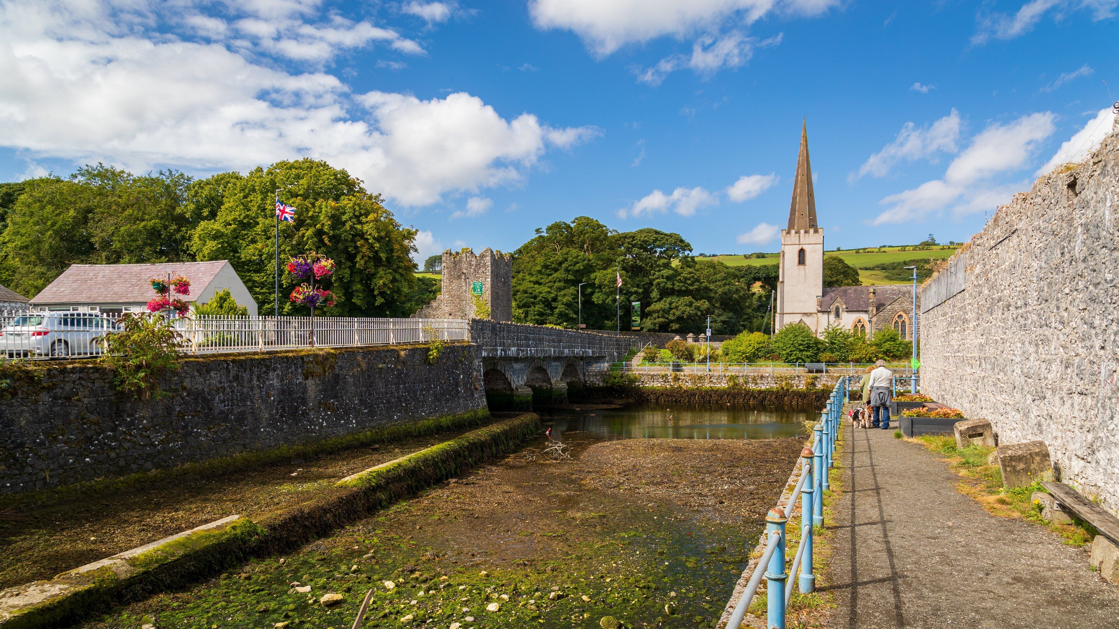 A stone bridge over a river, a church with a tall spire, and a white fence with flowers.
