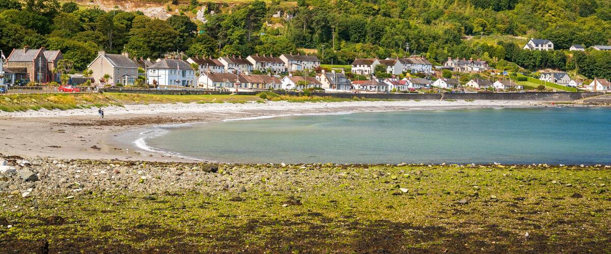 A coastal town with houses along the beach, a clear blue sea, and green hills in the background.