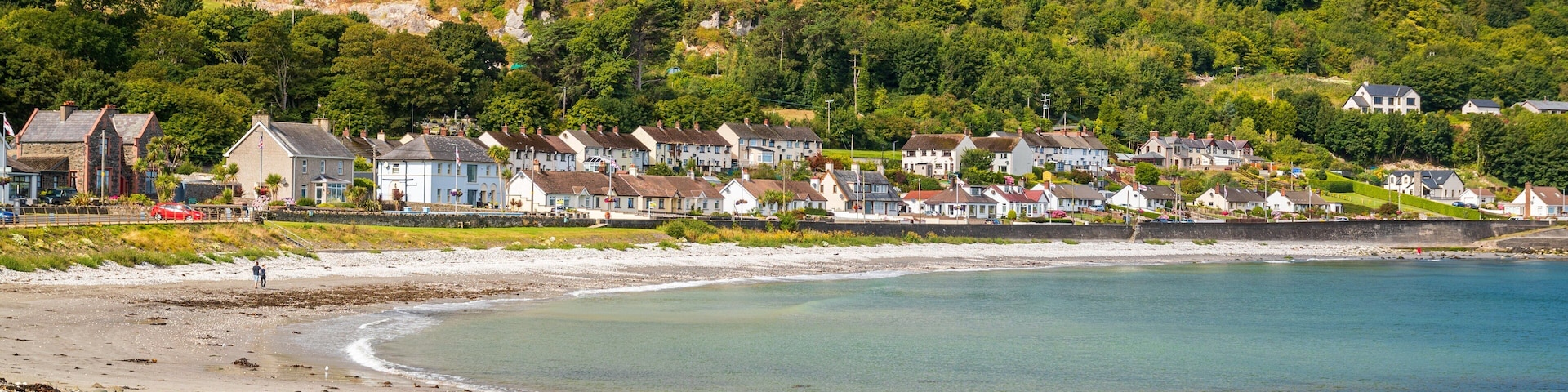 A coastal town with houses along the beach, a clear blue sea, and green hills in the background.