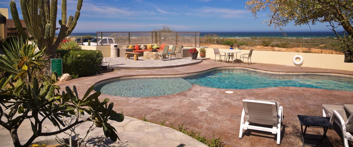 A swimming pool with a view of the ocean, surrounded by outdoor furniture and cacti.