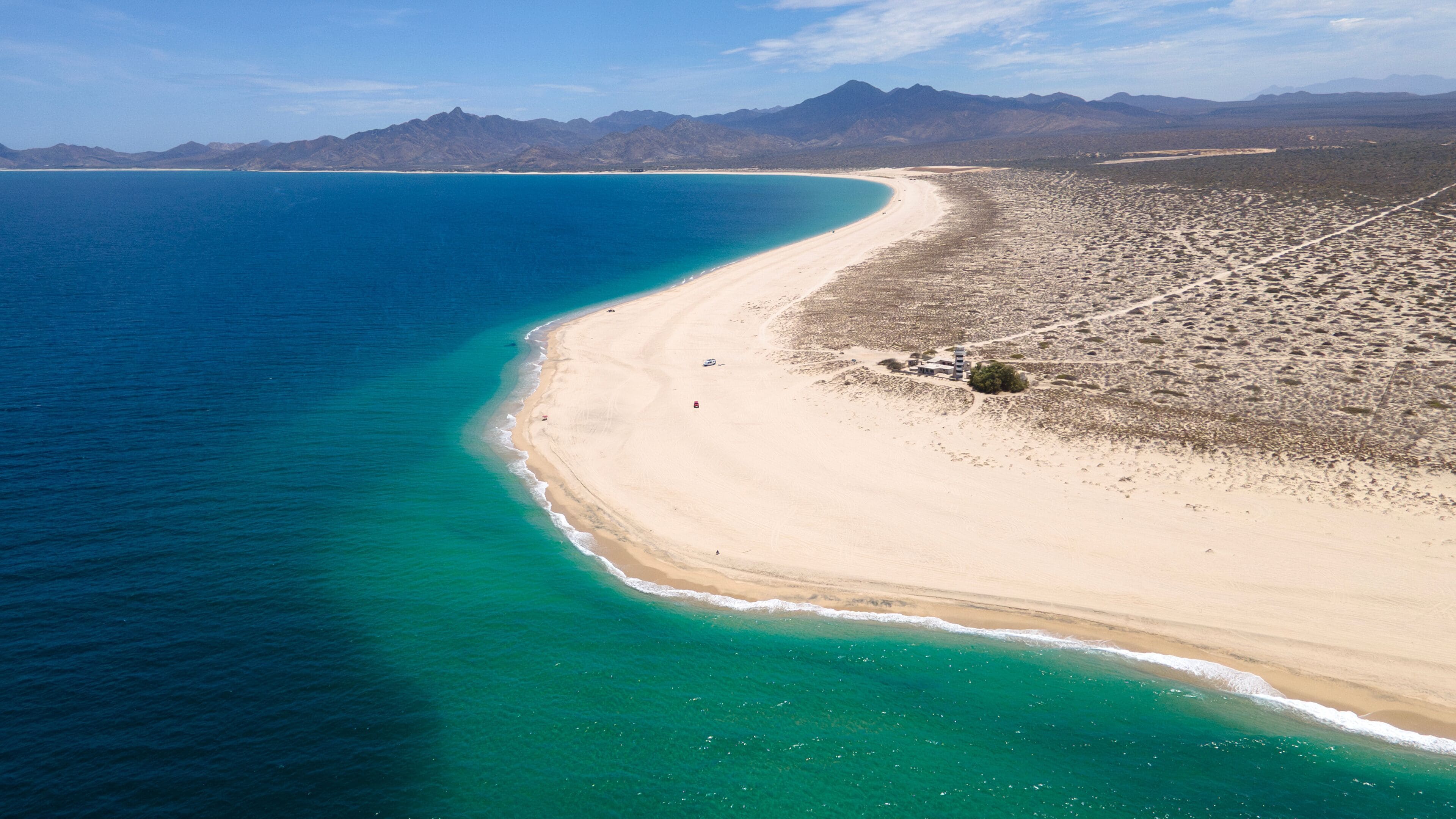 A coastal landscape with a sandy beach, clear turquoise waters, and distant mountains.