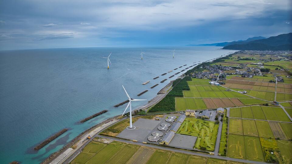 An aerial view of a coastal area with wind turbines, a harbor with breakwaters, and agricultural fields.