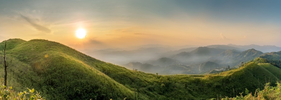 Un paisaje montañoso con colinas verdes y exuberantes, y un sol poniéndose al fondo.
