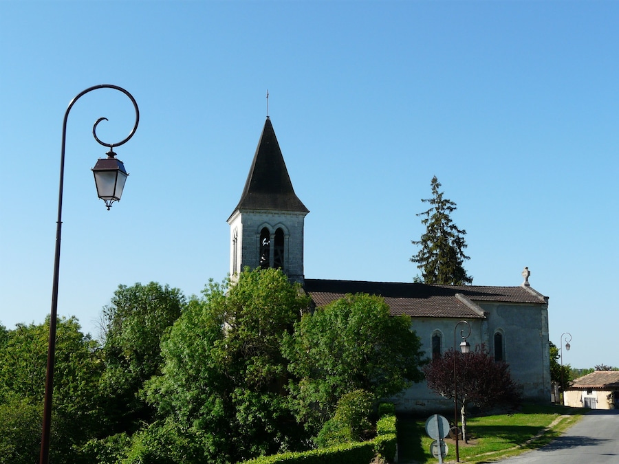 Une église dotée d’un clocher imposant et d’un beffroi, entourée d’arbres verdoyants et sous un ciel d’un bleu limpide.