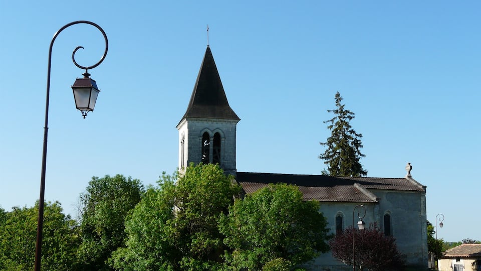 A church with a tall steeple and a bell tower, surrounded by lush green trees and a clear blue sky.