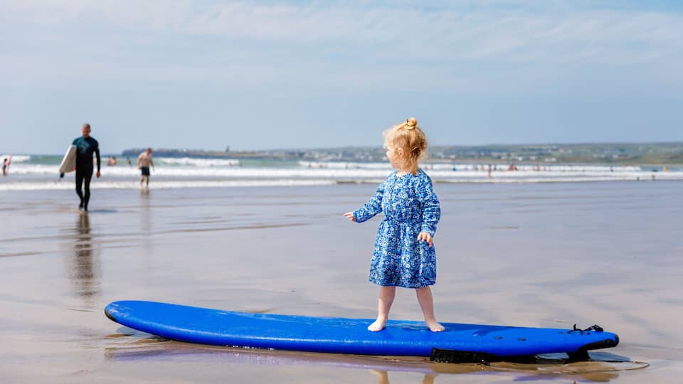 A child standing on a blue surfboard on a beach with waves in the background.