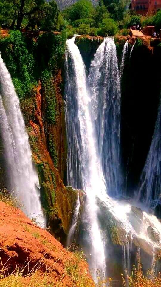 A multi-tiered waterfall with lush greenery and a red rock formation at the base.