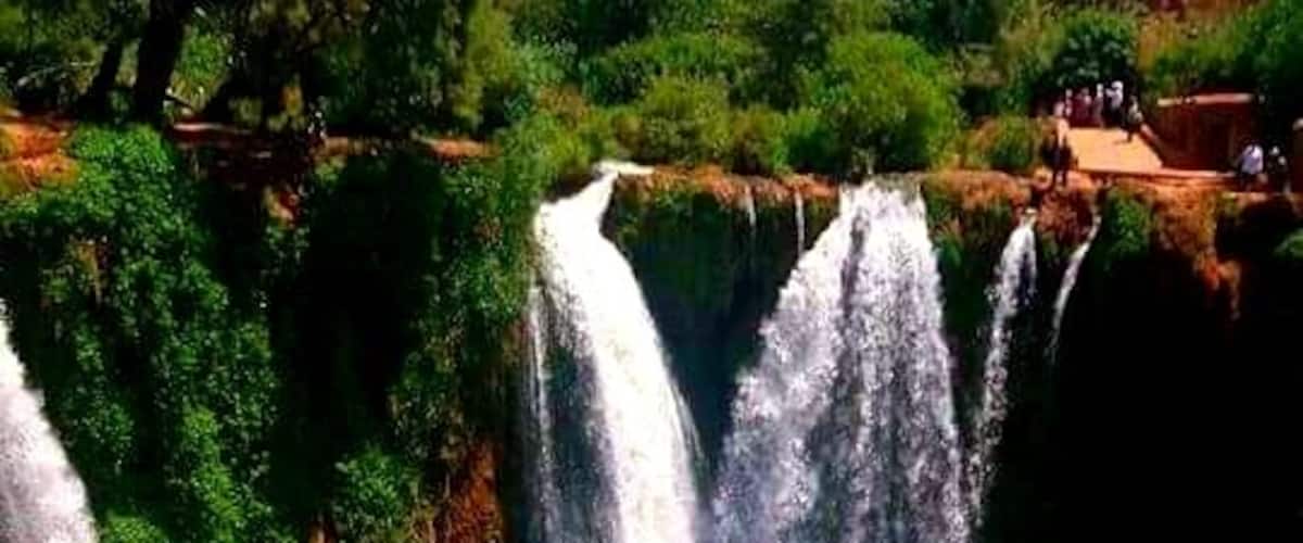 A multi-tiered waterfall with lush greenery and a red rock formation at the base.