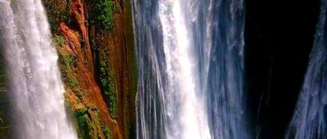 A multi-tiered waterfall with lush greenery and a red rock formation at the base.