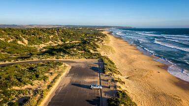 A coastal road with a parked car, sandy beach, ocean waves, and sparse vegetation.