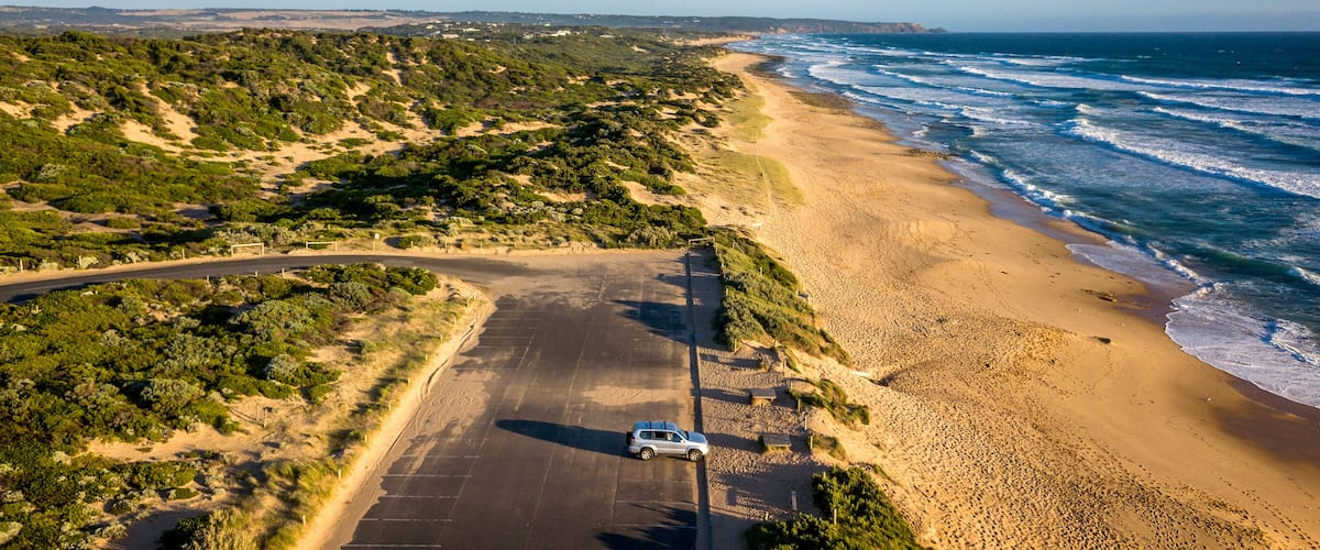 A coastal road with a parked car, sandy beach, ocean waves, and sparse vegetation.