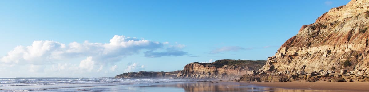 A beach with calm waters, sandy shore, and cliffs in the background.