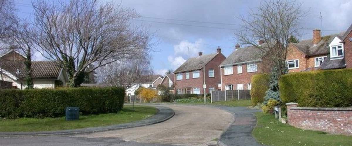 A residential street with houses, a curved road, and a grassy area with a hedge.