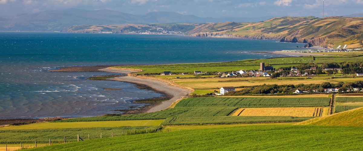 A coastal landscape with green fields, a beach, and distant hills.