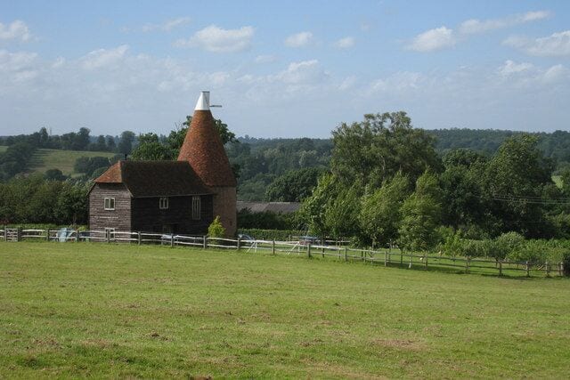 A traditional thatched-roof house with a conical roof, surrounded by a white fence, set in a lush green field with trees and hills in the background.