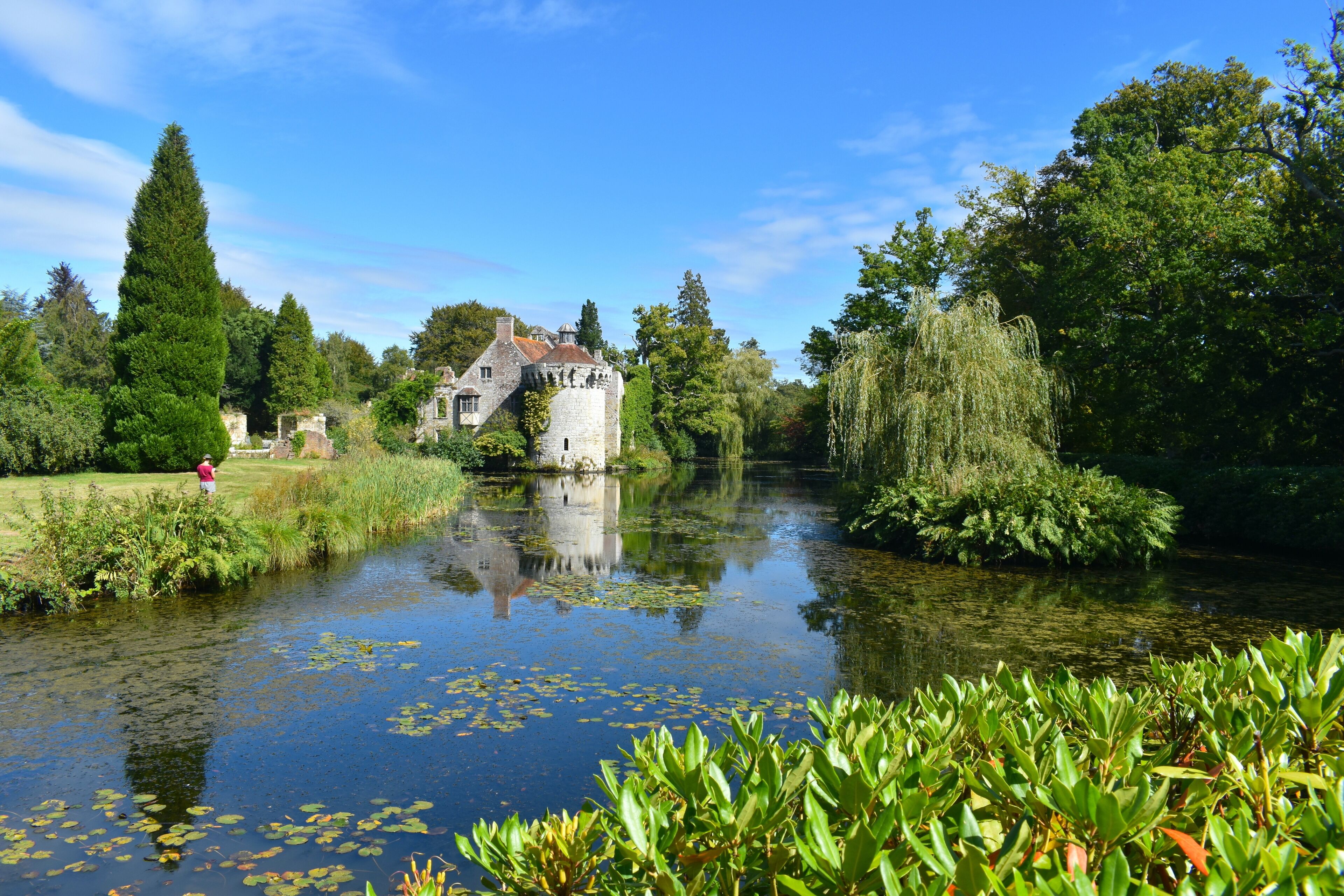 A silhouette of a woman drawing a British medieval castle and its reflection in water. Romantic gardens with panoramic views of a moated fortress and a Victorian country mansion all in a wooded estate