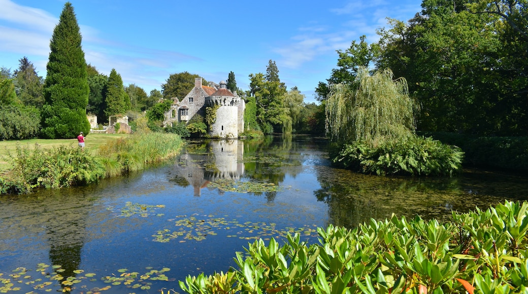 A silhouette of a woman drawing a British medieval castle and its reflection in water. Romantic gardens with panoramic views of a moated fortress and a Victorian country mansion all in a wooded estate