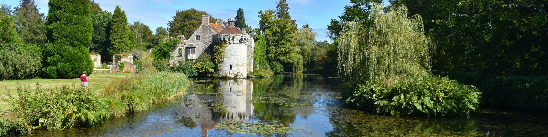 Escena tranquila junto al río con una torre de piedra histórica, abundante vegetación y una masa de agua en calma que refleja el entorno.