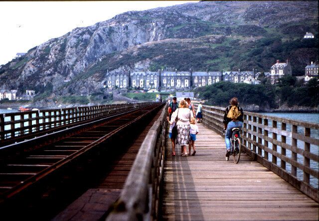 Uma ponte de madeira com pessoas caminhando e andando de bicicleta, com vista para um litoral rochoso e edifícios.