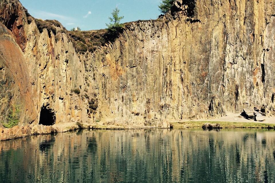 A clear lake reflecting a rocky cliff with a cave entrance.