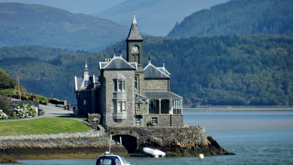 A historic stone building with a clock tower by a calm lake, surrounded by lush green hills.