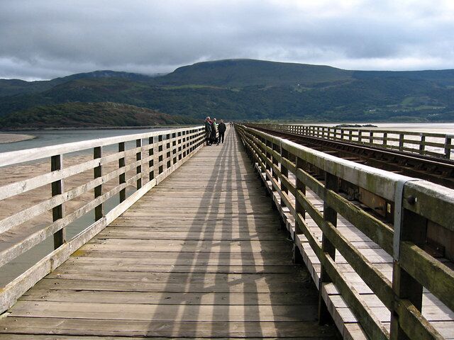 A wooden bridge with railings, leading to a body of water and mountains in the background.