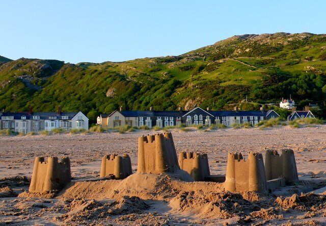 A sandy beach with sand castles, a row of houses, and a hill with greenery in the background.