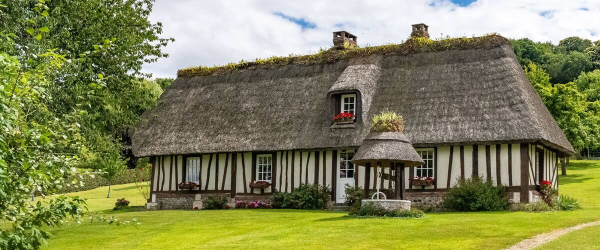 A thatched-roof cottage with a small window and a door, surrounded by a well-maintained lawn and trees.