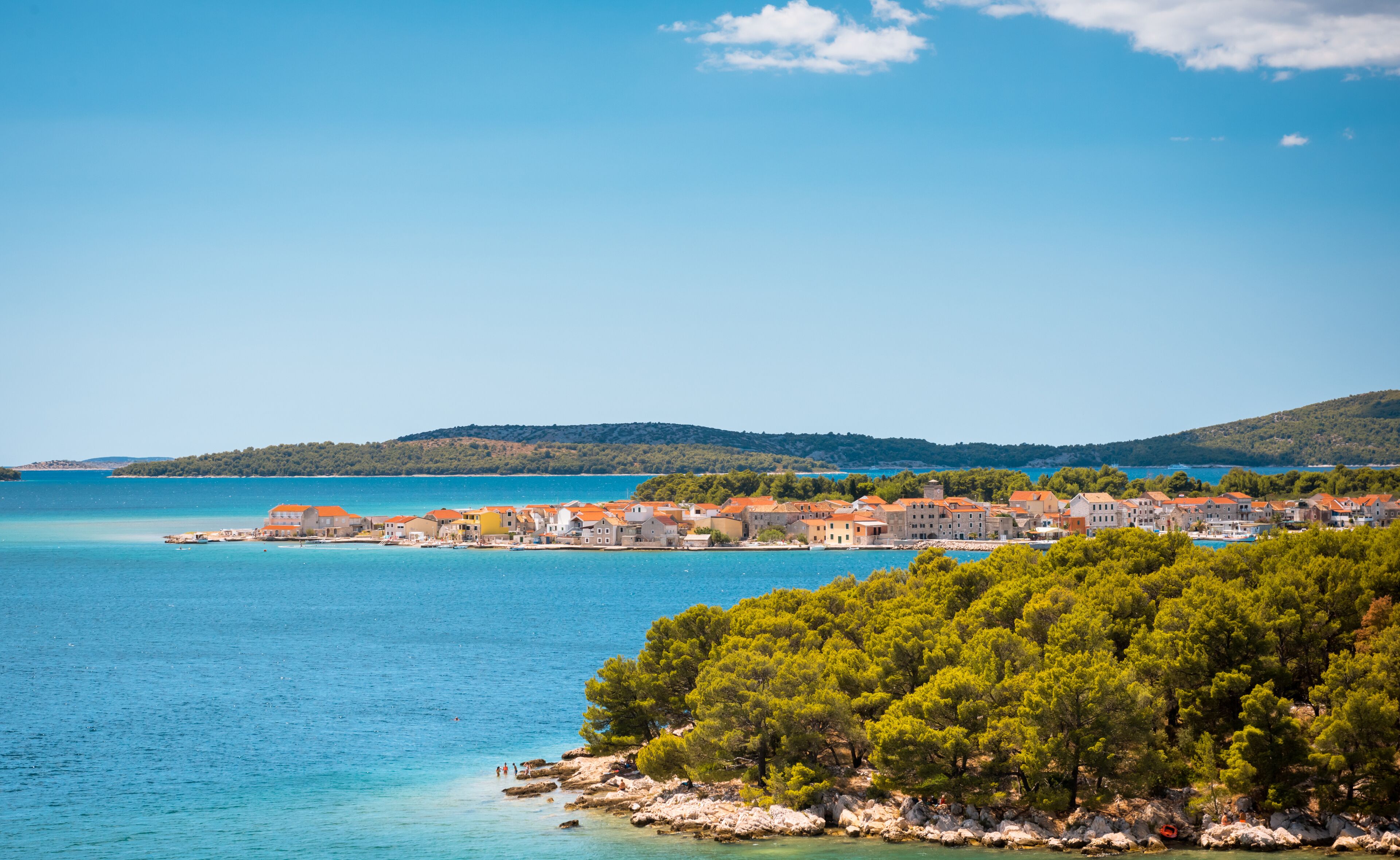 A coastal town with buildings along the shore, a clear blue sea, and a forested hill in the background.