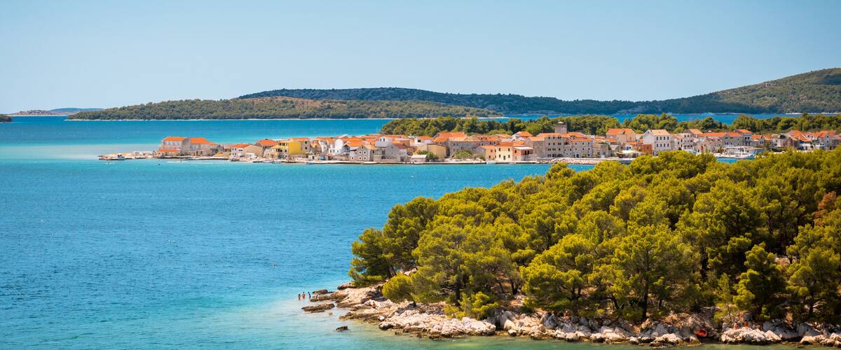 A coastal town with buildings along the shore, a clear blue sea, and a forested hill in the background.