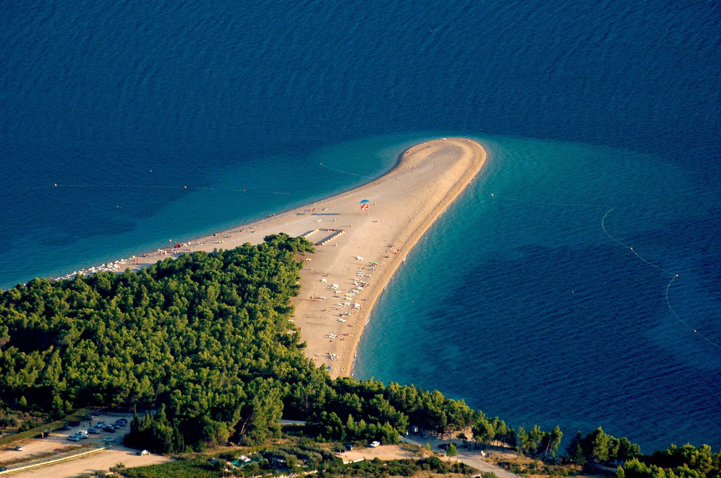Luchtbeeld van een kustgebied met een gebogen strand, dichtbegroeid bos en helderblauw water.