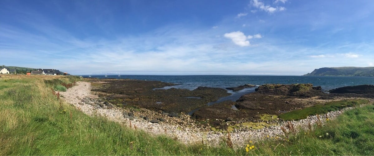 A coastal landscape with rocky shores, grassy areas, and distant hills under a clear sky.