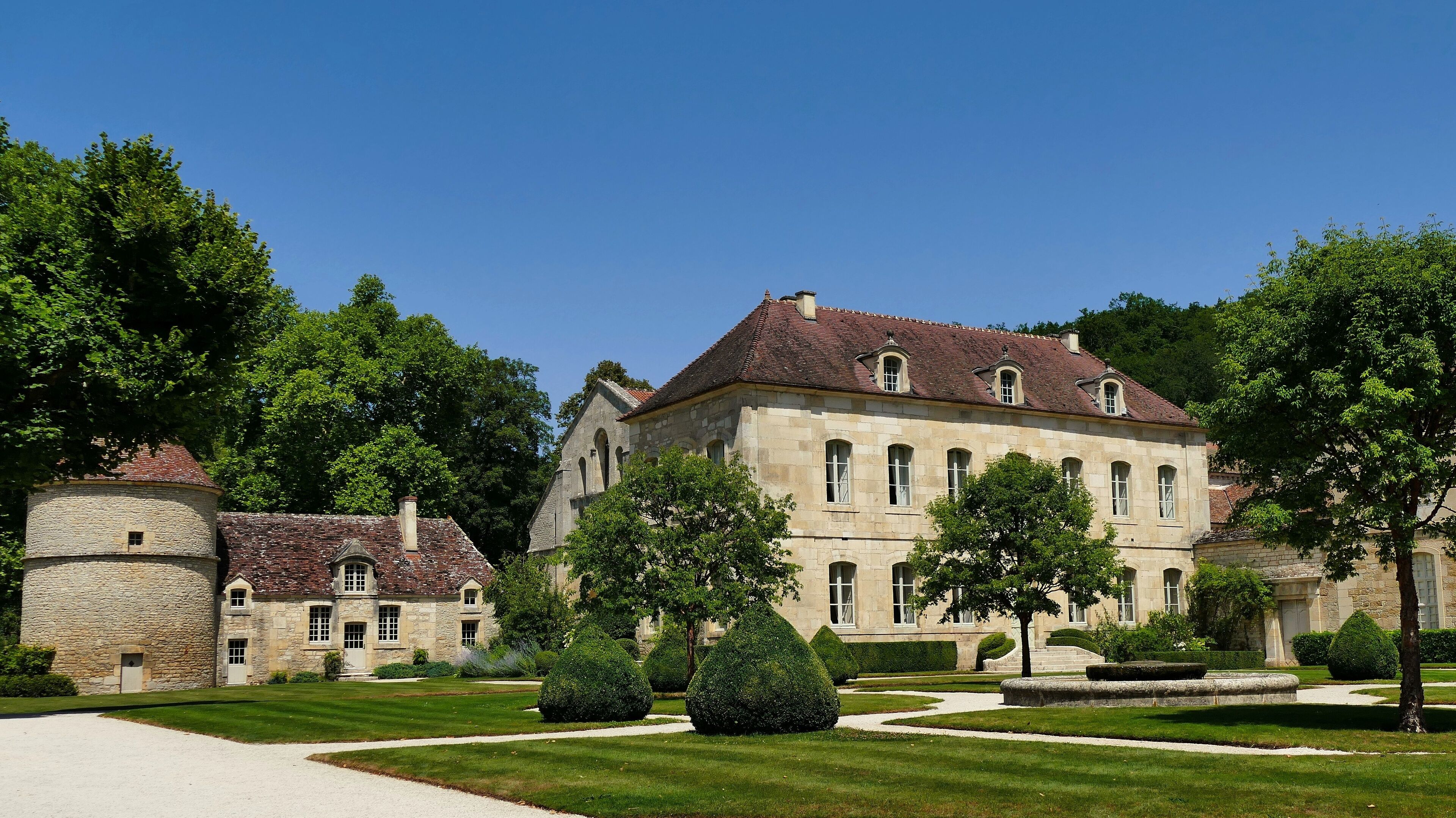 A historic stone building with a red-tiled roof, surrounded by well-manicured lawns and trees.