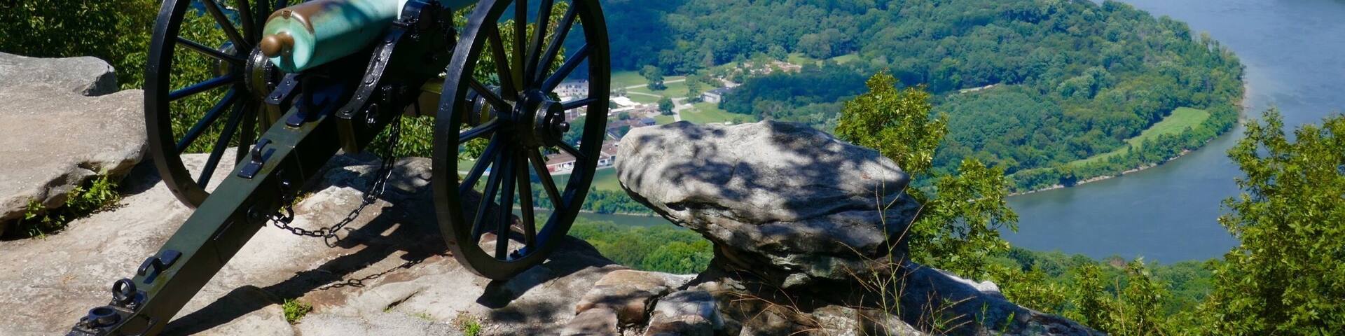 A cannon on a rocky outcrop overlooking a river and city.