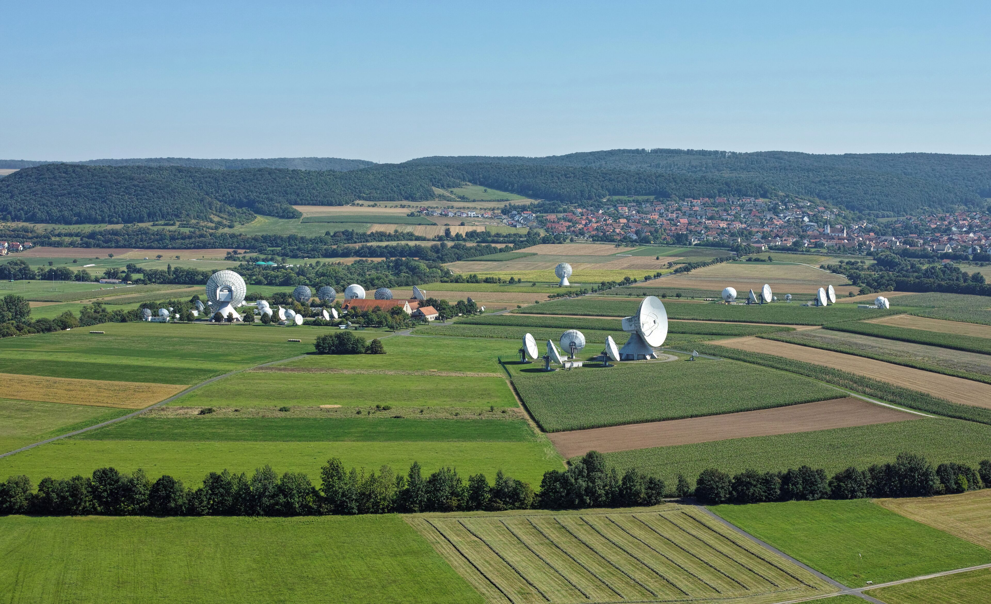 A rural landscape with agricultural fields, a village in the distance, and multiple large satellite dishes.