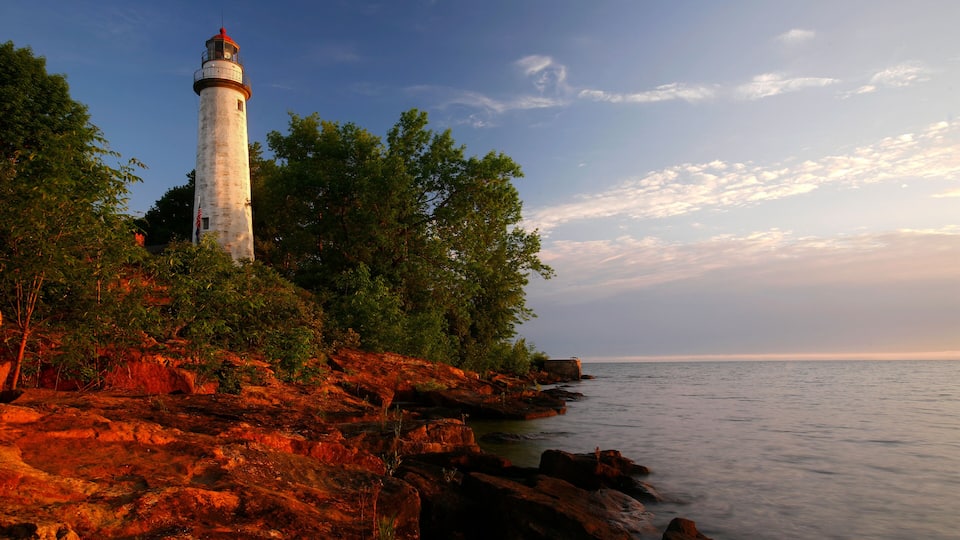 A lighthouse on a rocky coastline with trees and calm sea.