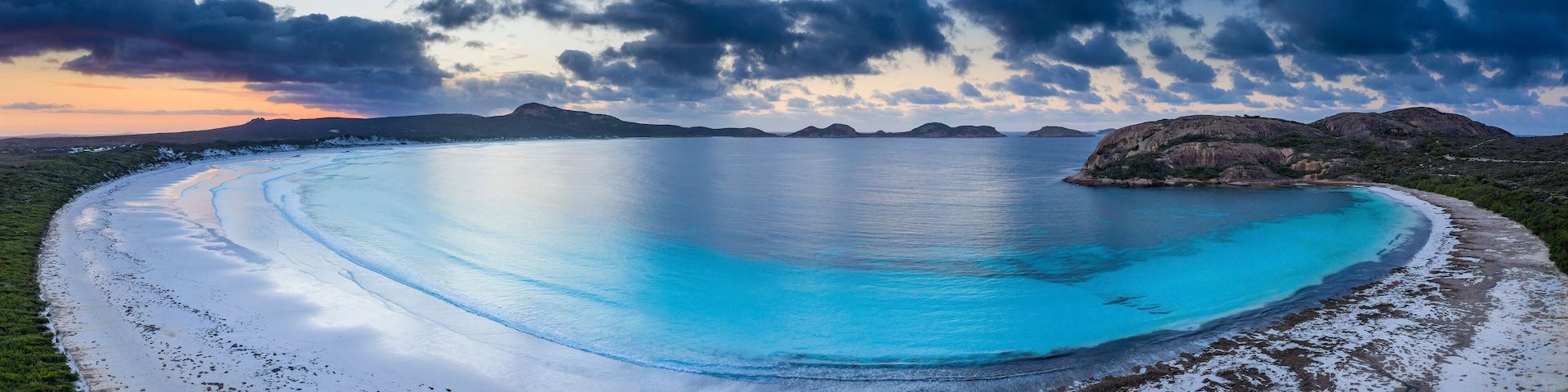 A coastal landscape with a clear blue sea, sandy beach, and rocky outcrops.