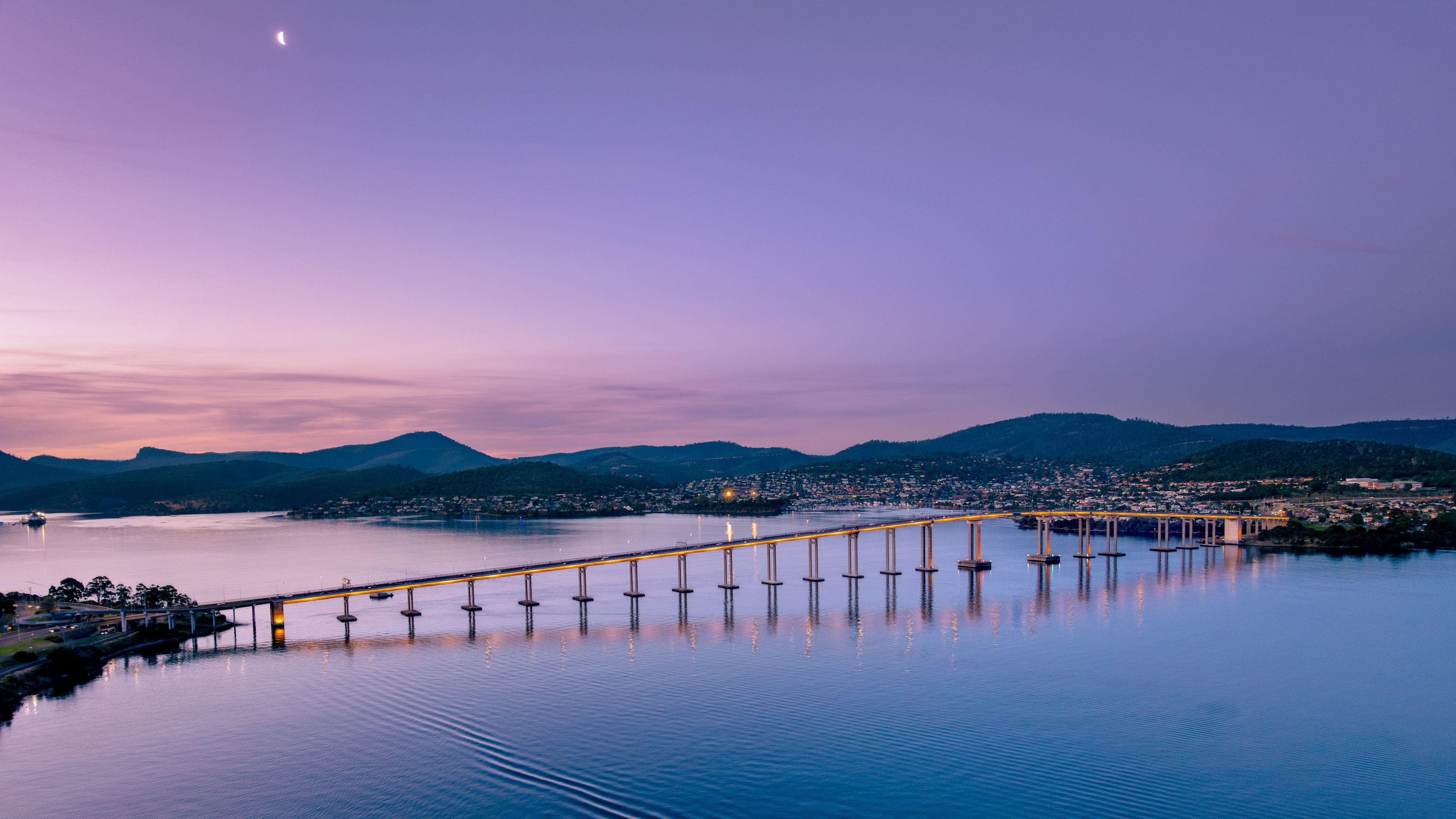 A bridge over calm water with a city in the background and mountains in the distance.