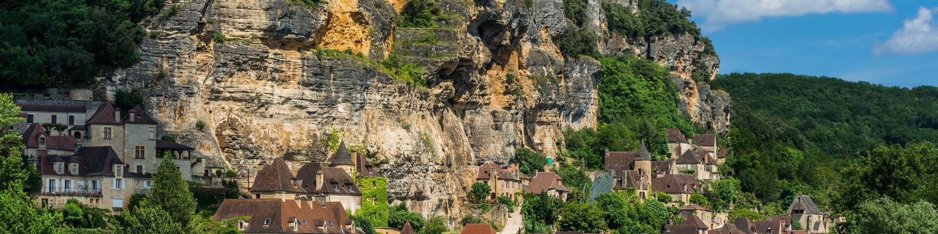 Un pueblo a orillas de un río, con edificios frente al agua, un acantilado destacado con una cueva y abundante vegetación.
