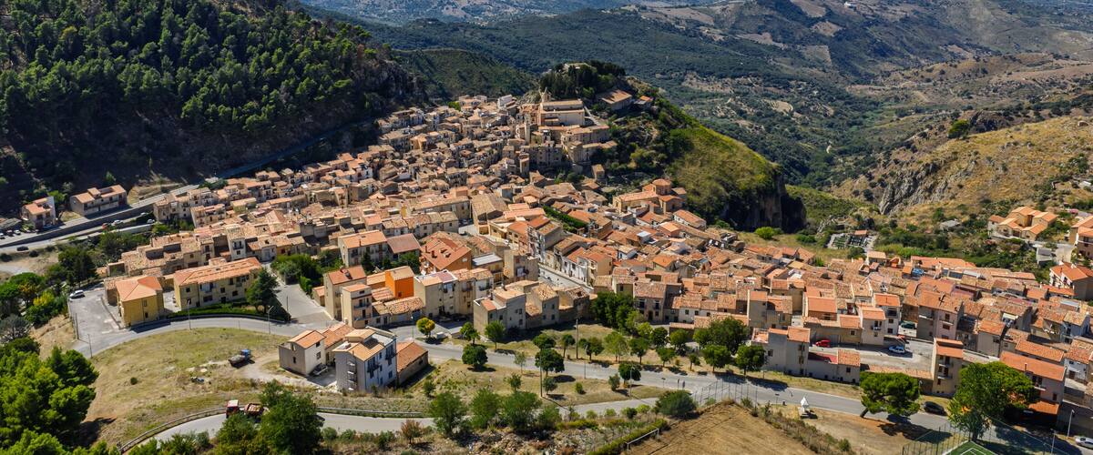 A coastal town with terracotta-roofed buildings nestled in a valley, surrounded by hills and mountains.