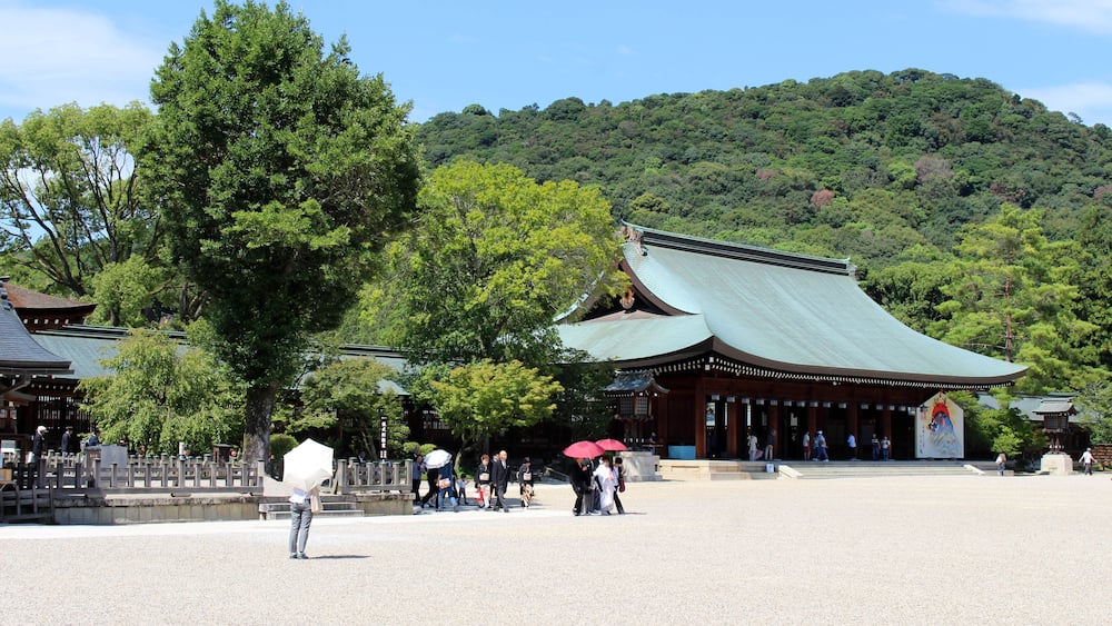 A traditional Japanese temple with a green roof, surrounded by lush greenery and a hilly backdrop.