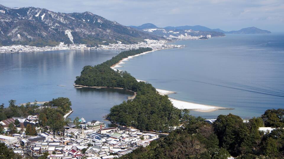 A coastal town with a curving bay, dense forest, and mountains in the background.