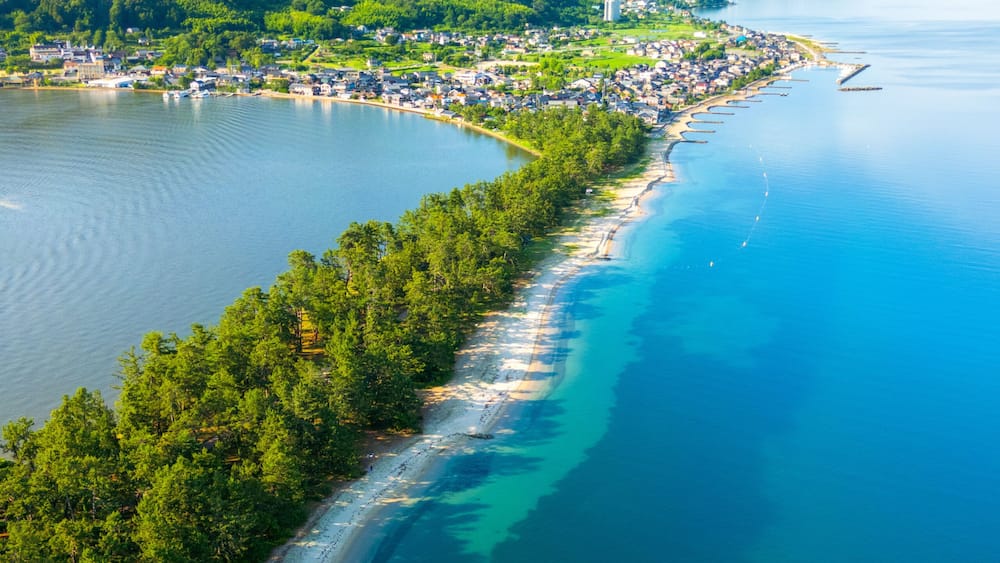 A coastal town with a long beach, dense forested hills, and a clear blue sea.