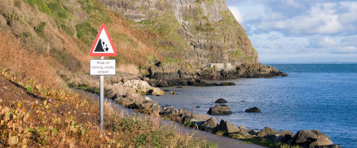 A coastal cliff with a warning sign for falling rocks, a lighthouse, and a rocky shoreline.
