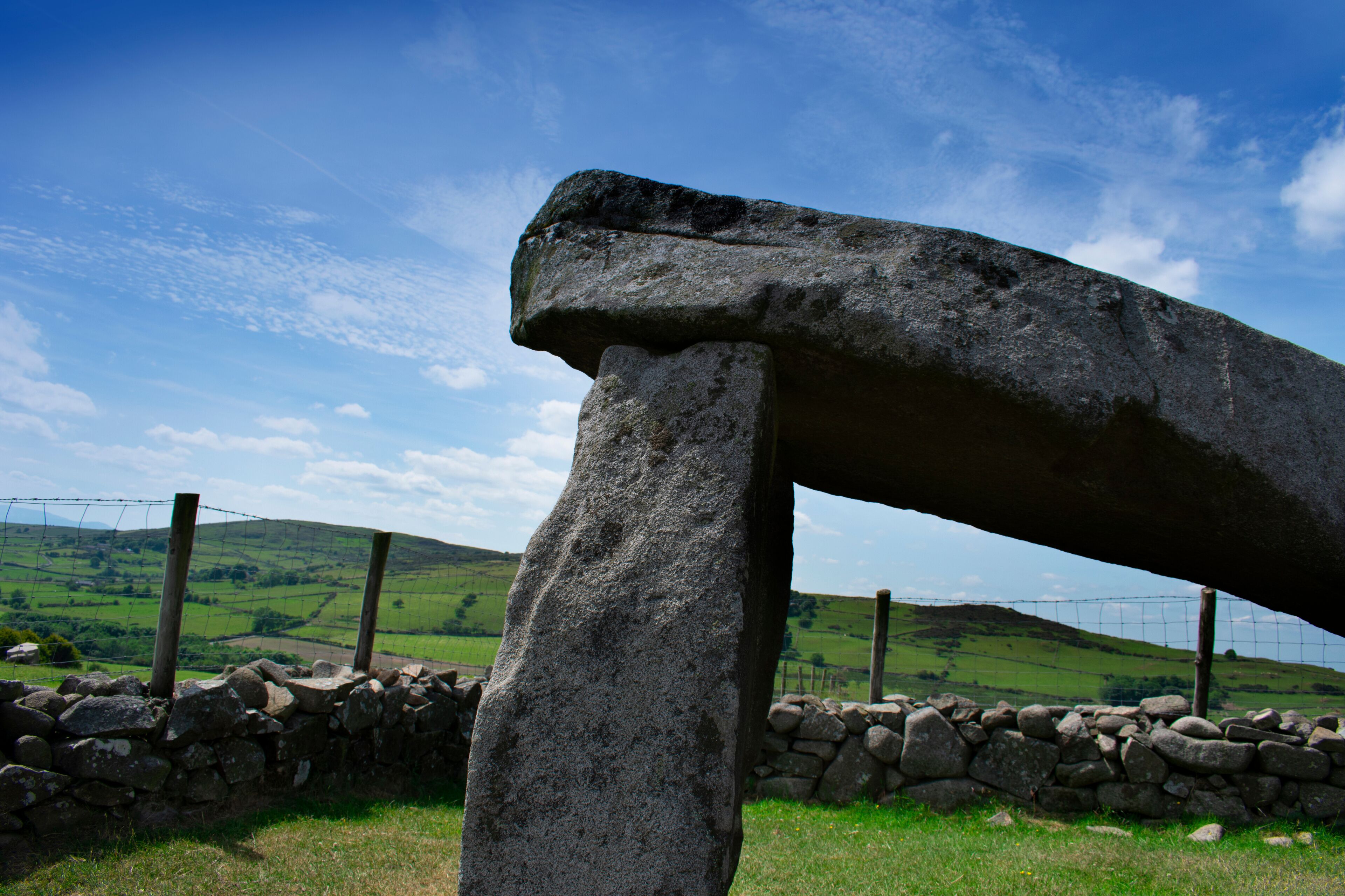A large stone structure in a rural landscape with a stone wall and a fence.