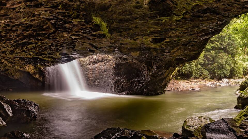 A waterfall in a cave with moss-covered rocks and a forested area beyond.