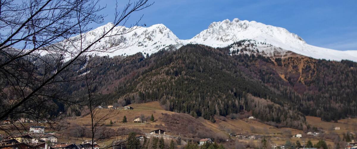 A town nestled at the base of snow-capped mountains, with a mix of residential buildings and a forested hillside.