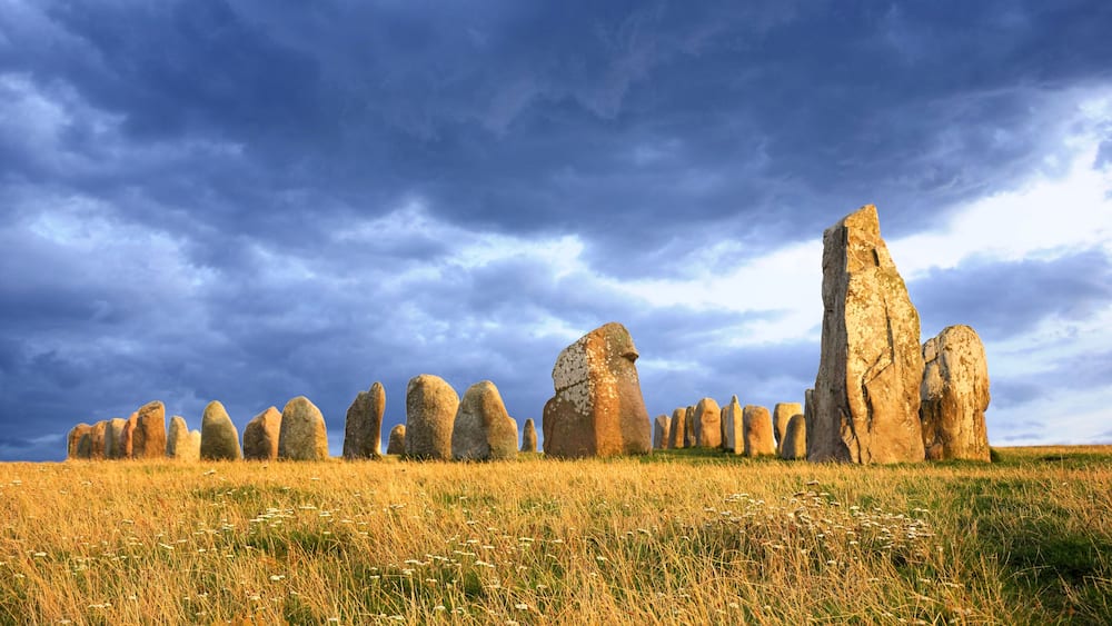 Ein Steinkreis mit hohen, aufrechten Steinen in einem grasbewachsen Feld unter einem bewölkten Himmel.